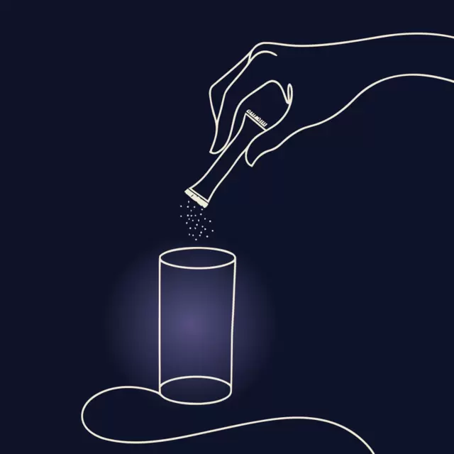 A hand tipping a single stick of powder into a tall glass of water that glows softly.